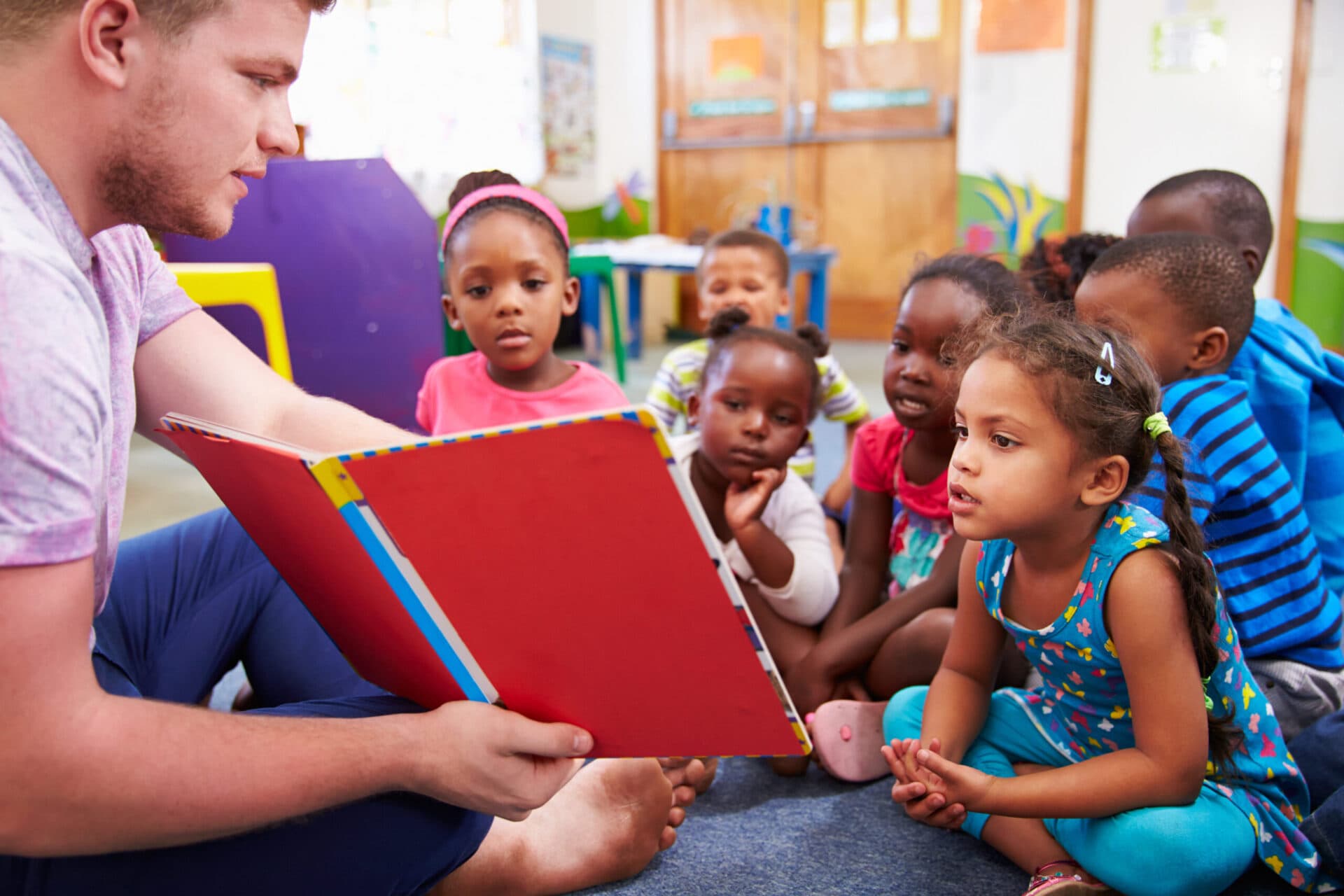 A teacher holding an open picture book while a group of children listen on the carpet