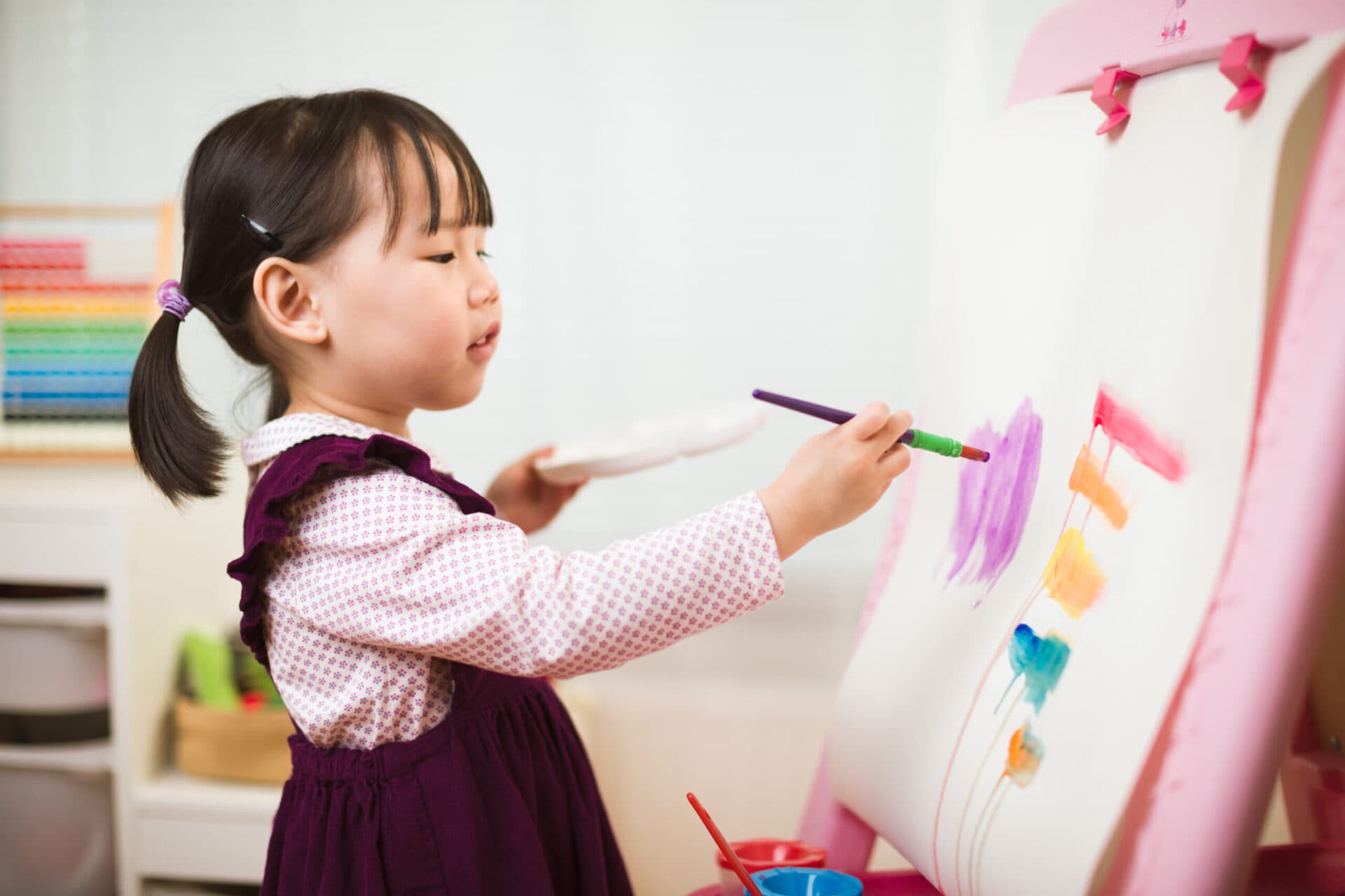 A young girl painting broad strokes of purple and orange on an easel