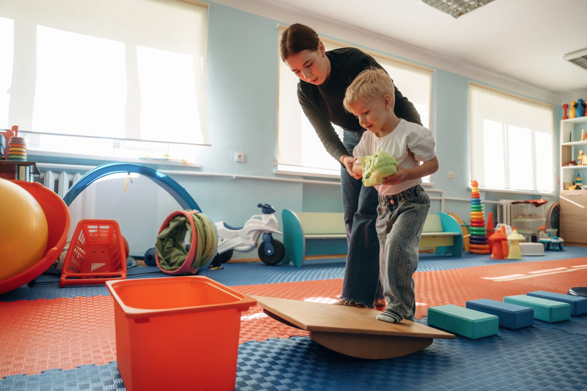 A teacher steadying a young child as he balances on a wooden wobble board