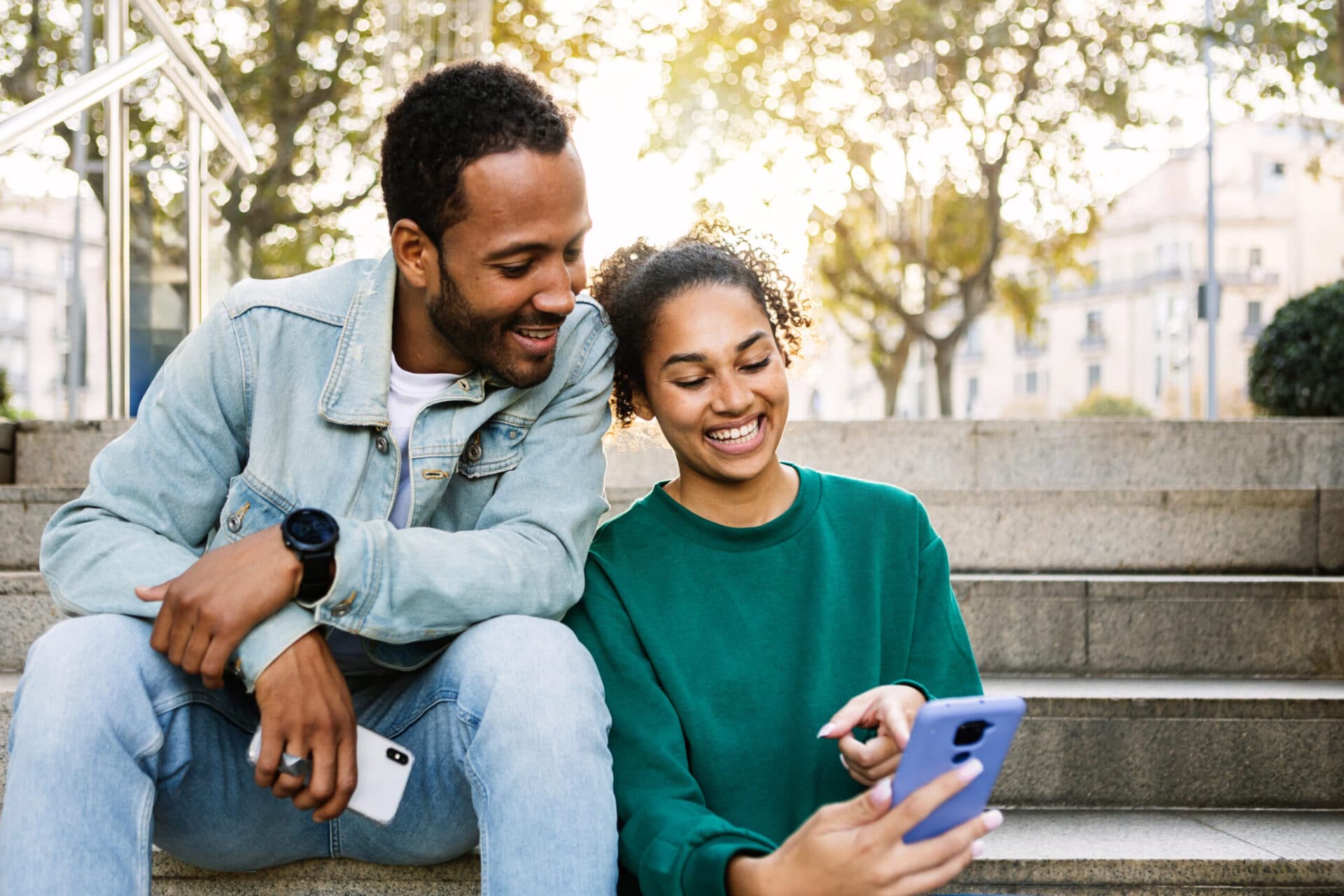 Two parents sitting outdoors on steps, smiling at updates on a phone
