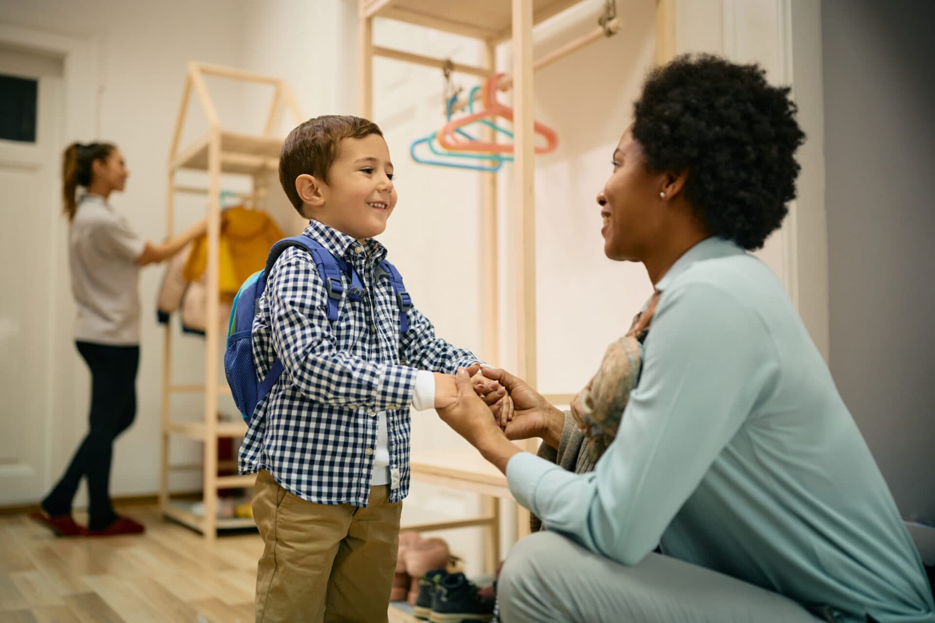 A teacher crouching to greet a smiling child wearing a backpack at the cubbies