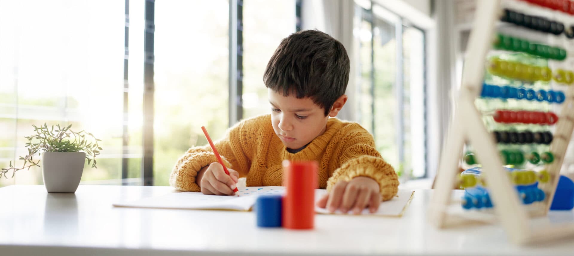 A boy in a yellow sweater concentrating as he writes in a workbook with a red pencil