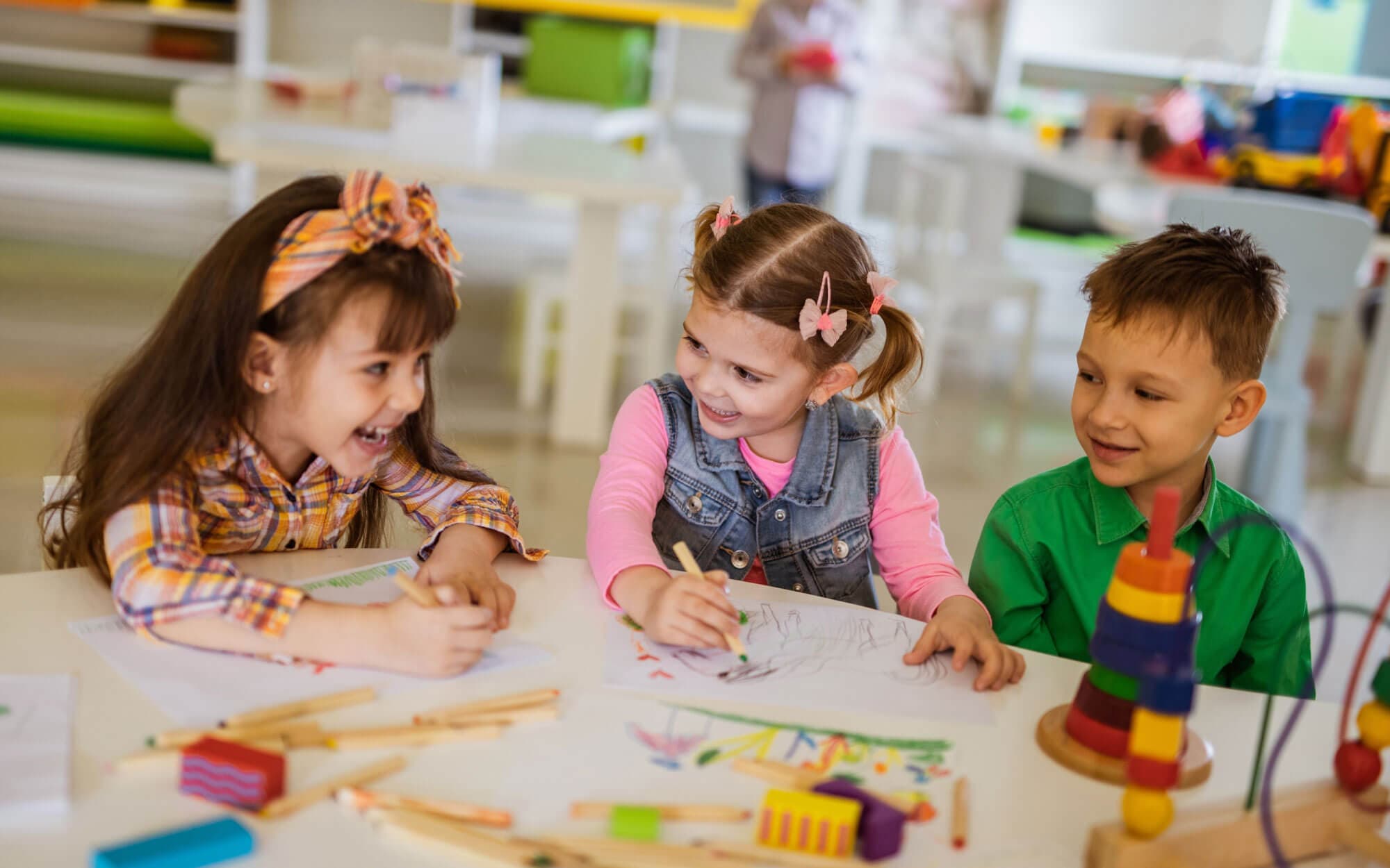 Three children laughing together while drawing with colored pencils at a table