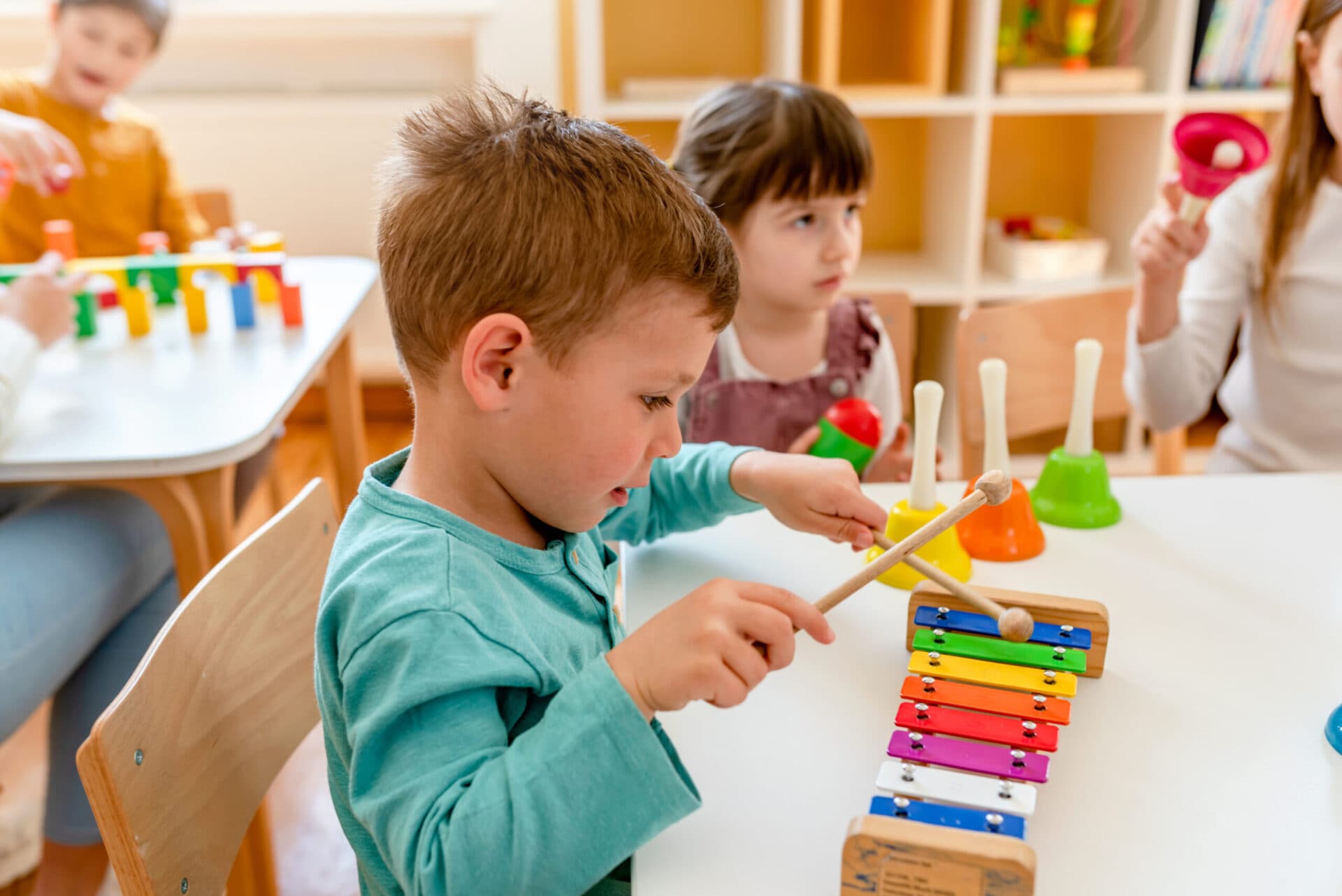 A child striking a rainbow xylophone with mallets at a table of hand bells