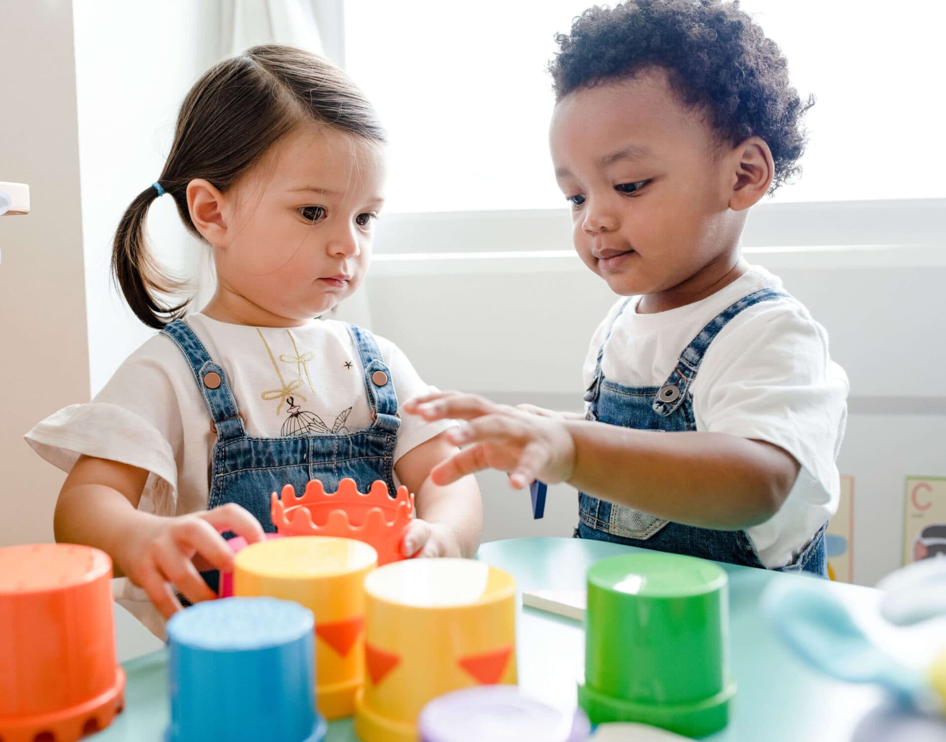Two toddlers at a table reaching for brightly colored stacking cups together