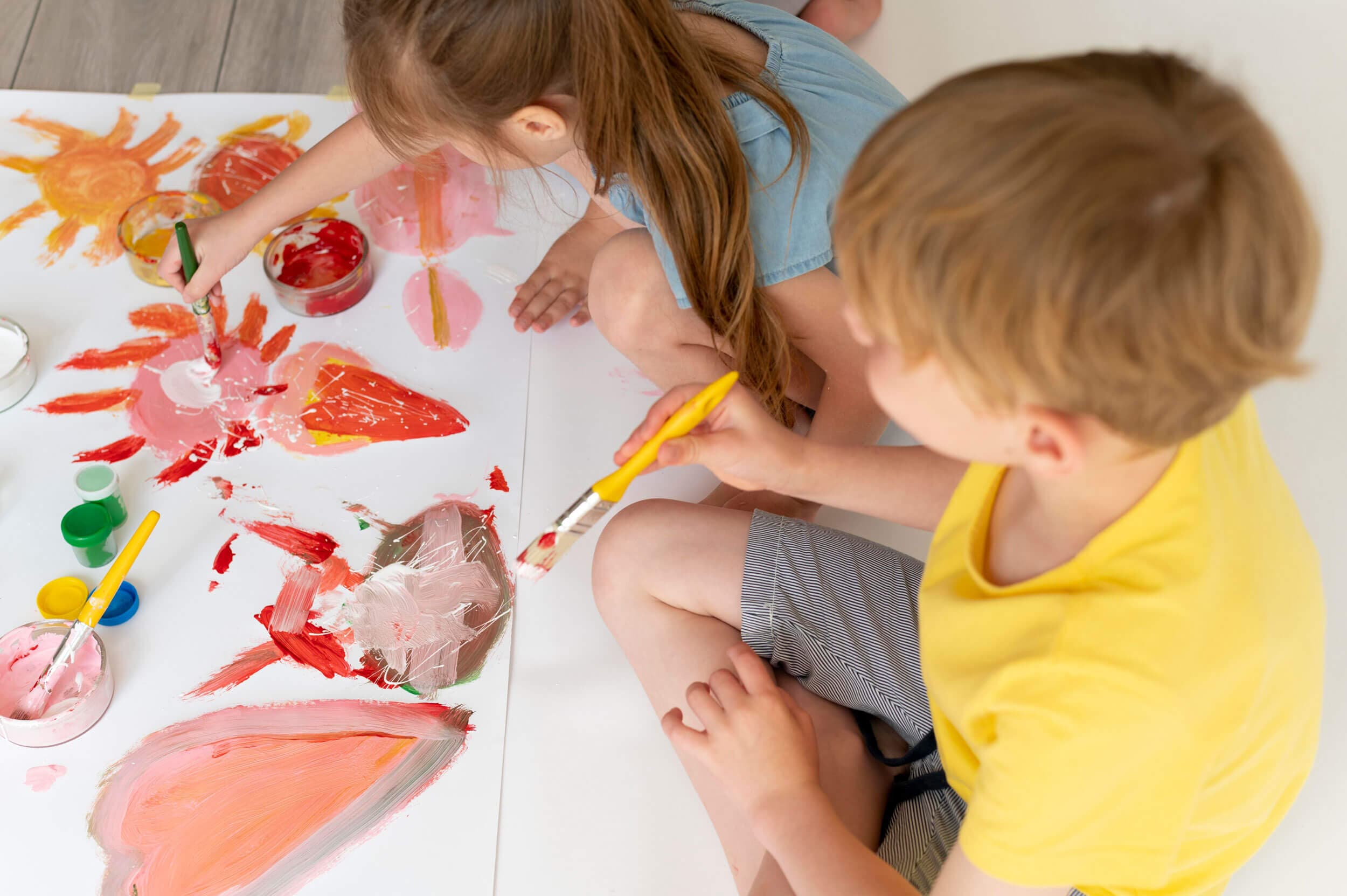 Two children kneeling on the floor painting a large sheet of paper with red and orange