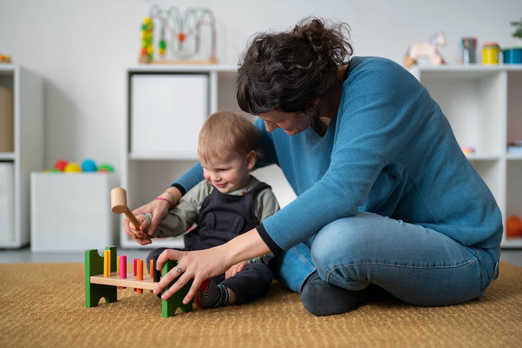 A caregiver sitting on the floor guiding a baby using a wooden hammer-and-peg toy