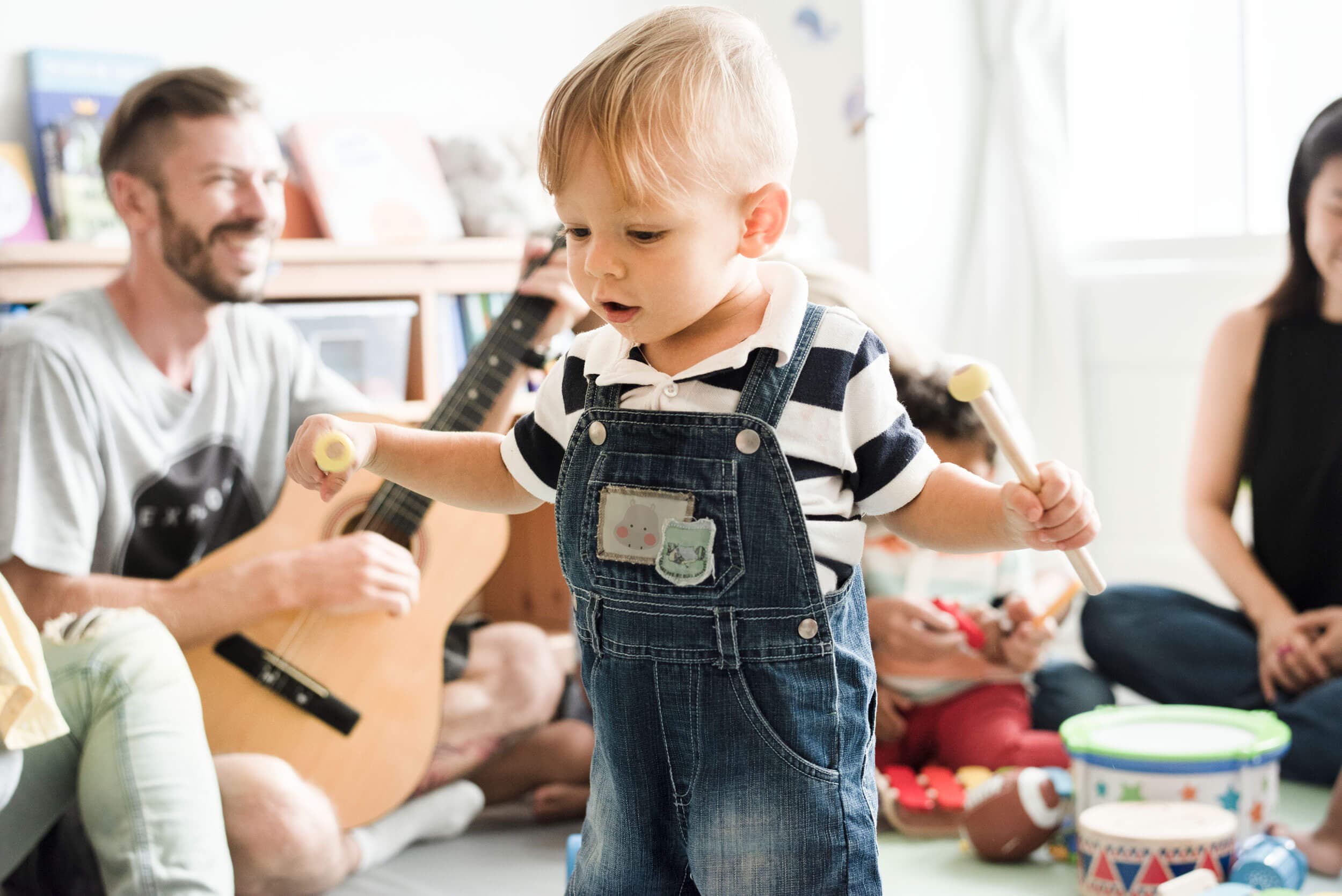 A toddler tapping rhythm sticks together during a music circle while a teacher plays guitar