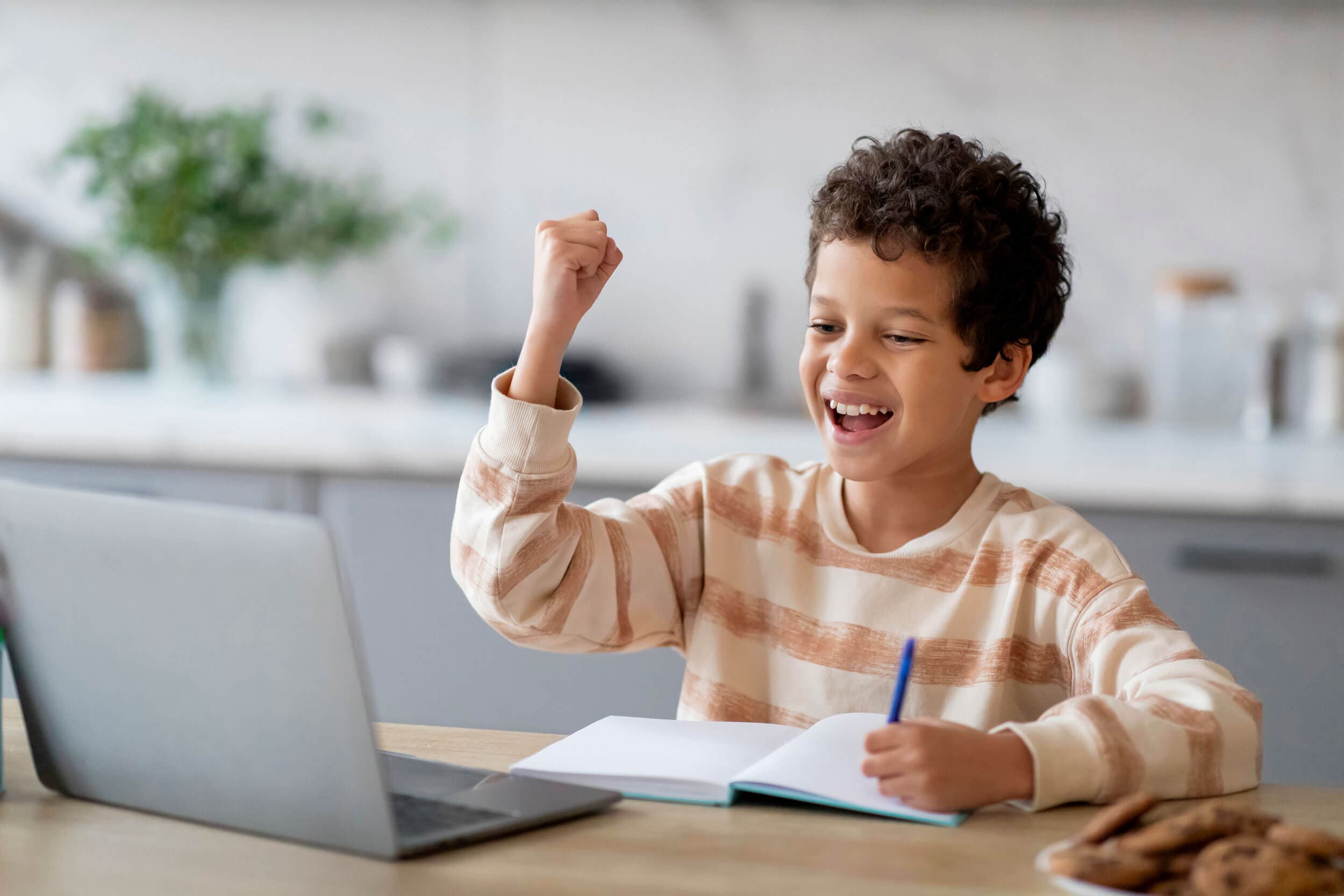 A boy pumping his fist and grinning at a laptop with an open notebook