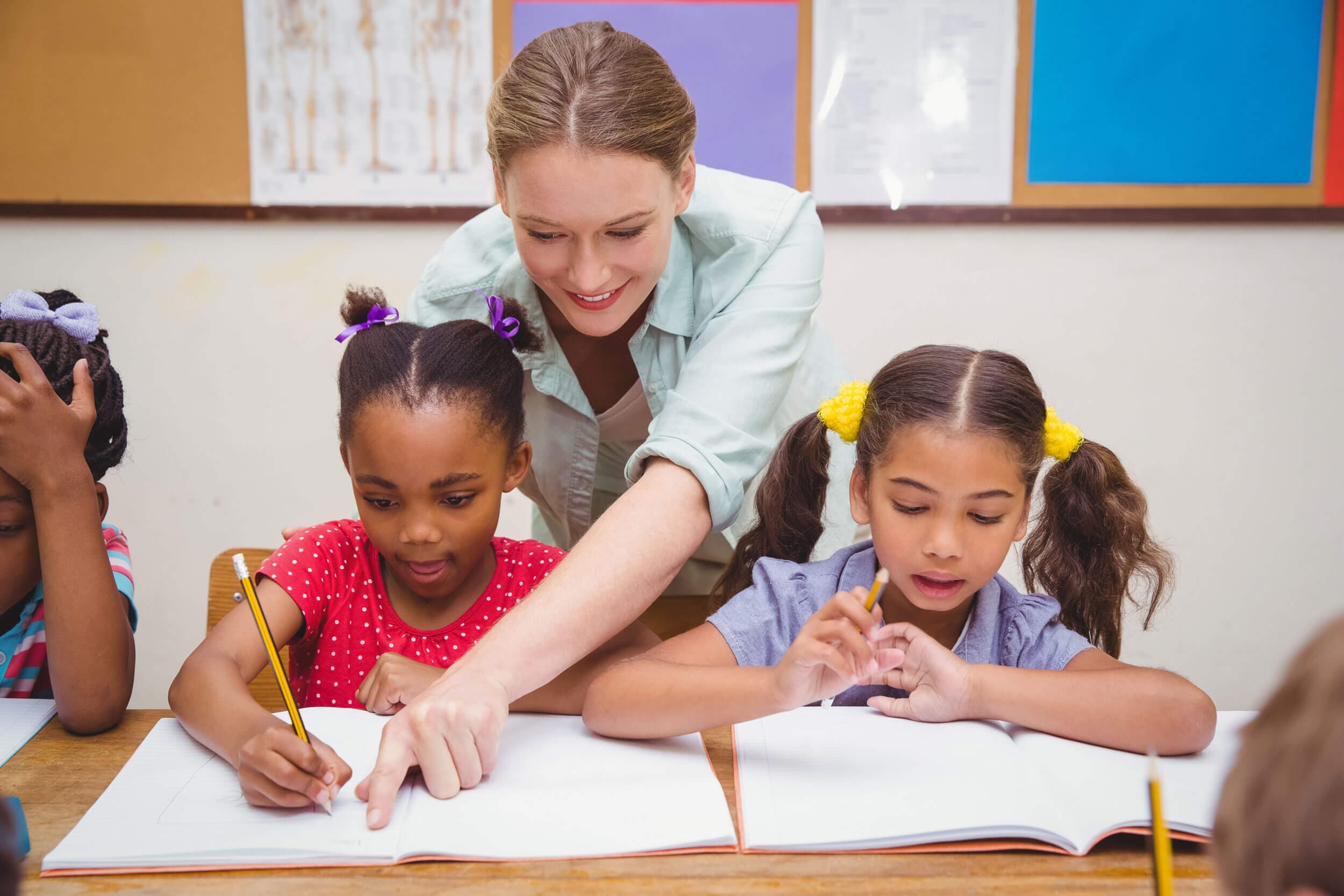 A teacher leaning in to point at a student's workbook while two girls write