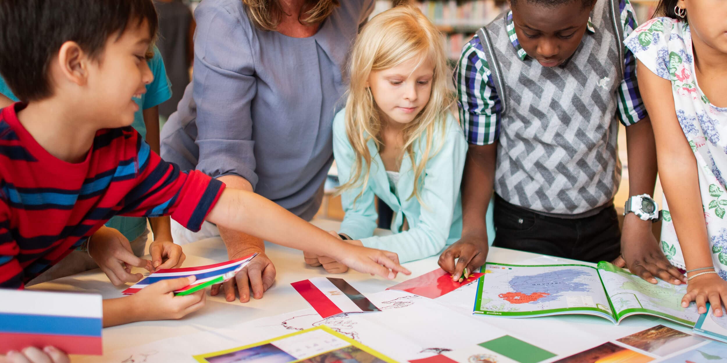 Children leaning over a table of world flags and an atlas with their teacher