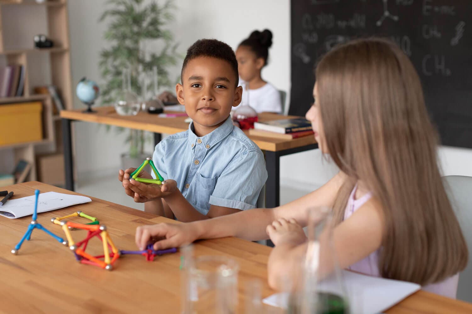 A boy holding up a geometric shape he built from magnetic rods beside a classmate