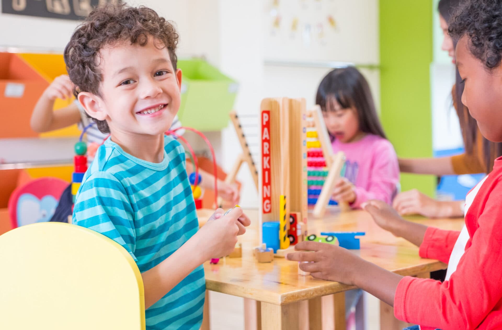 A smiling preschooler at a table of wooden toys, turning to face the camera