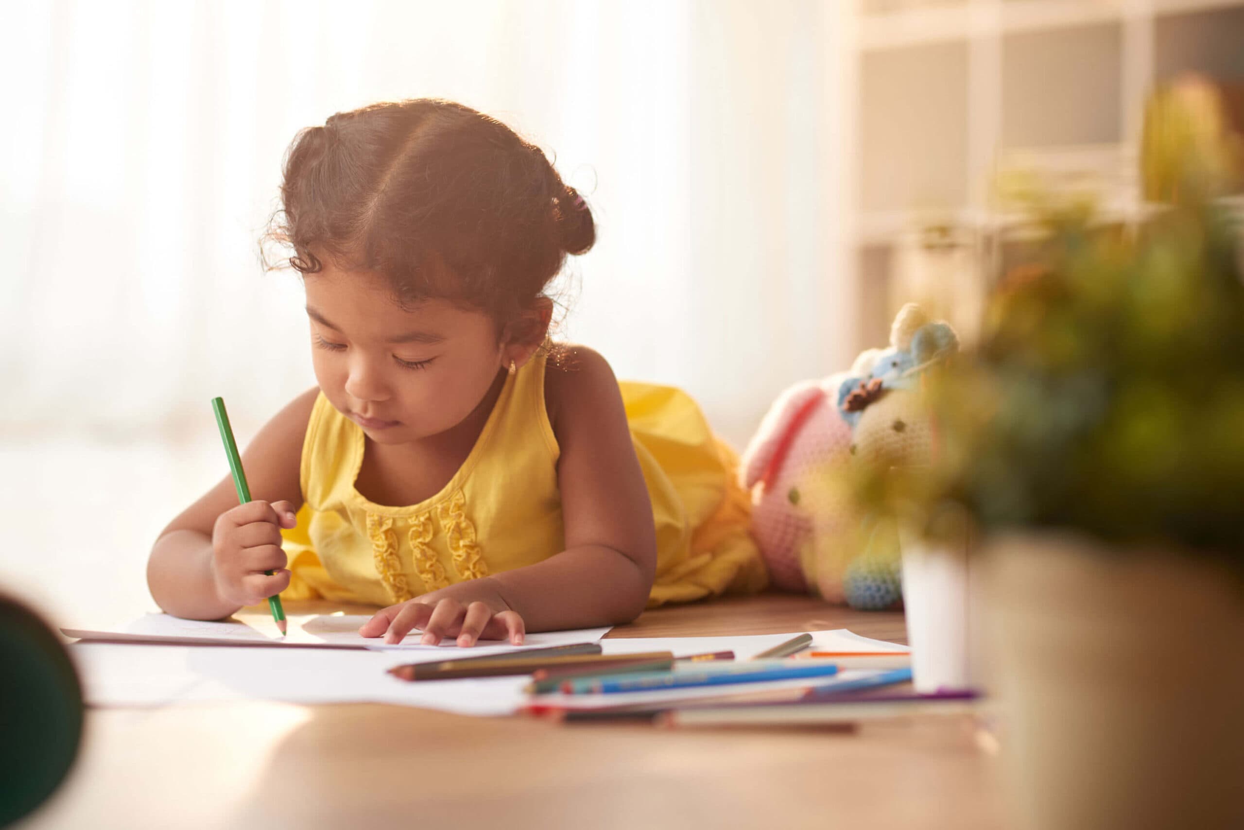 A young girl lying on the floor drawing with a green colored pencil on paper