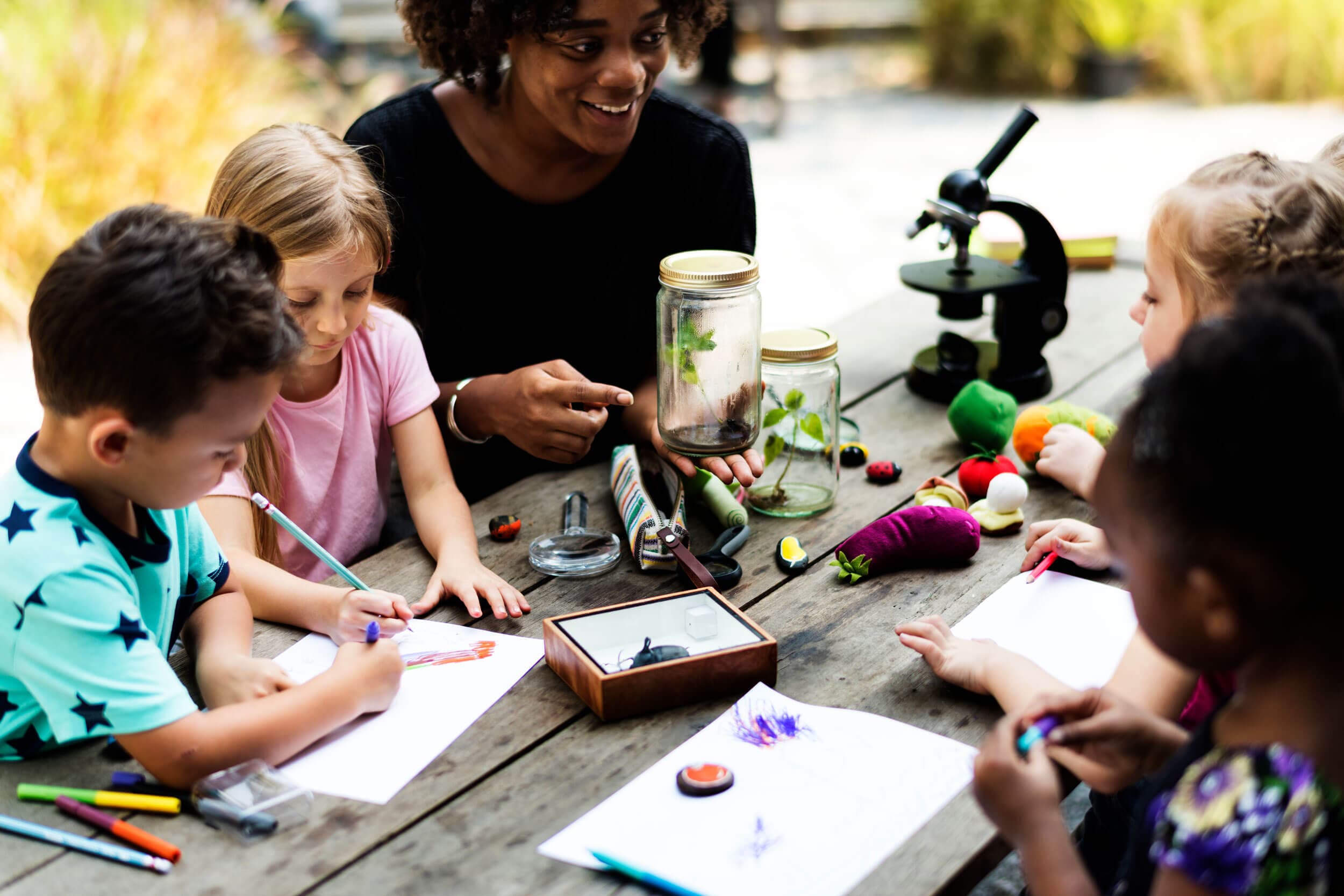 A teacher and children examining insect specimens in jars at an outdoor picnic table