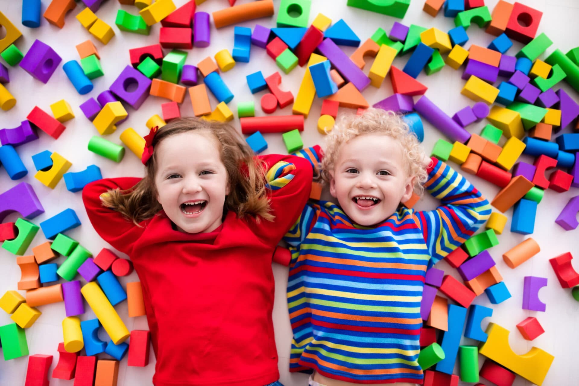 Two children lying side by side and laughing, surrounded by scattered colorful foam blocks