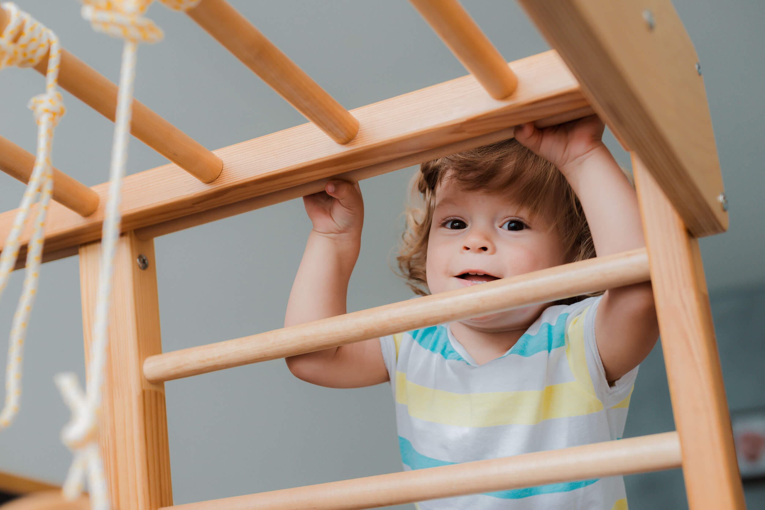 A smiling toddler gripping the rungs of a wooden climbing frame