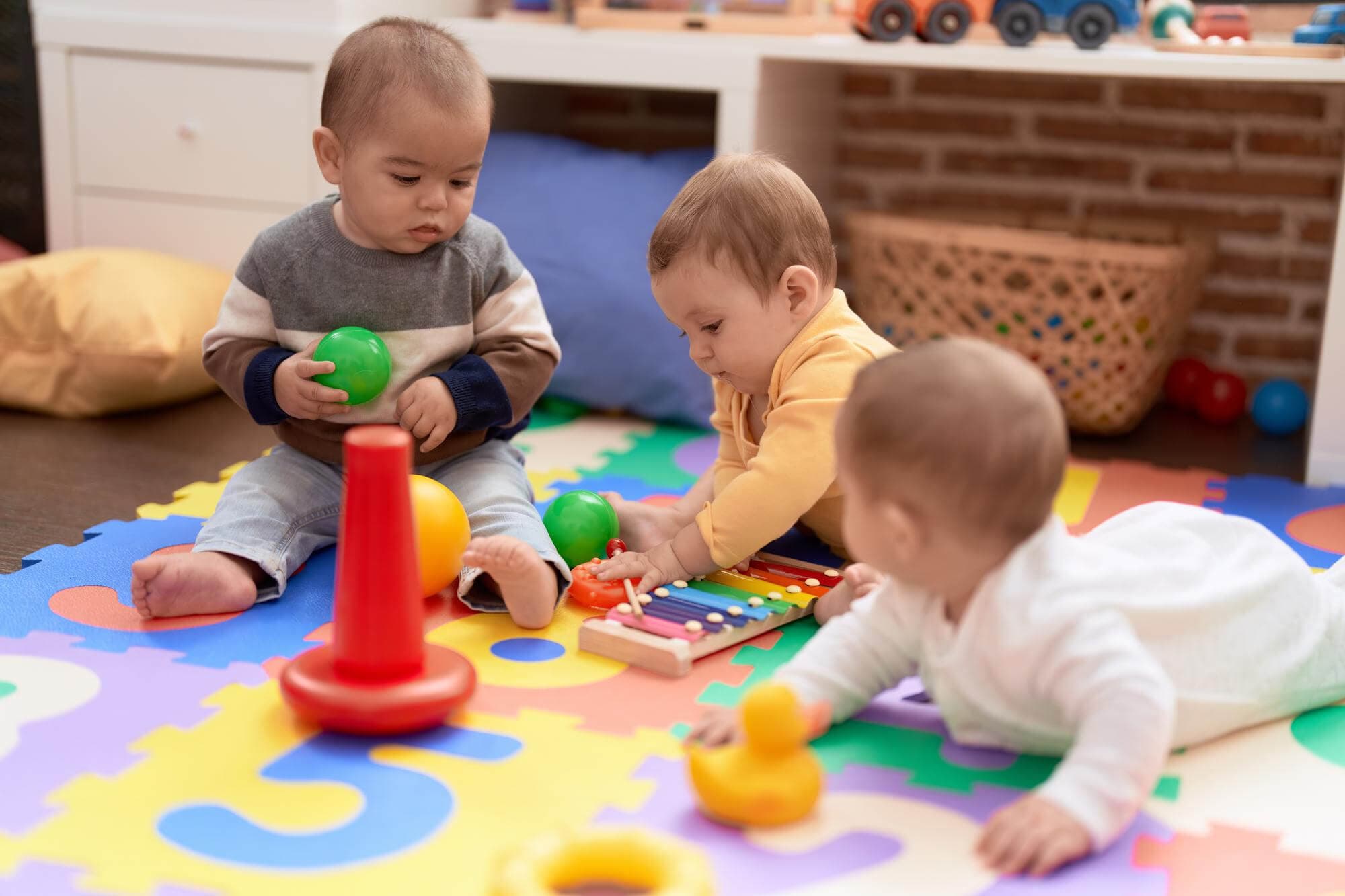 Three babies on a play mat exploring balls, a stacking toy and a xylophone