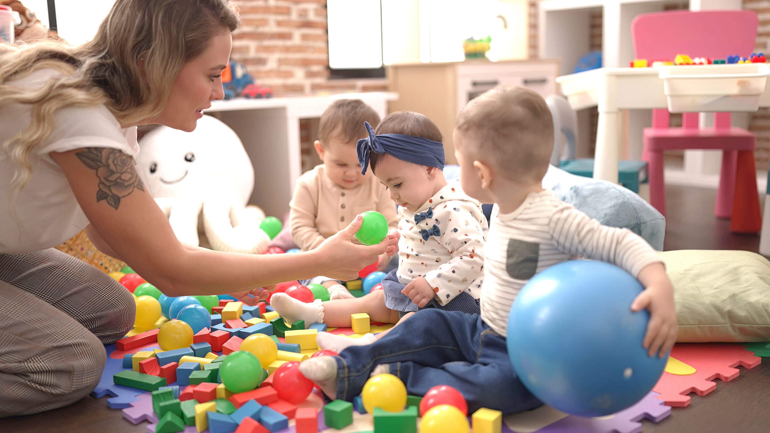 A teacher kneeling on the floor offering a green ball to three toddlers playing among building blocks