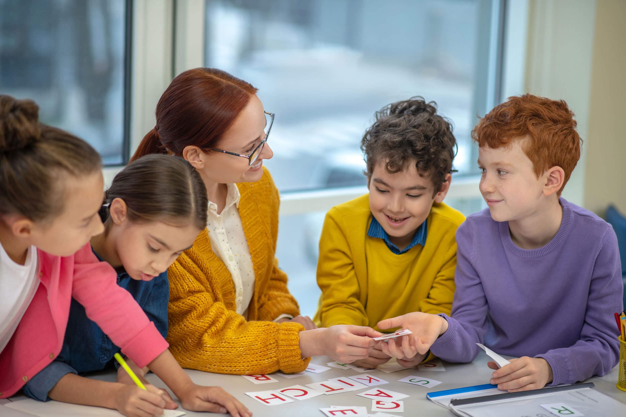 A teacher handing out letter cards to four children spelling words at a table